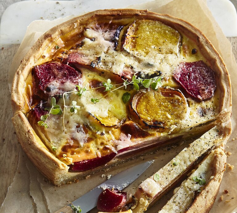 Angeschnittene Quiche mit Thymianzweig, geschmolzenem Käse auf Backpapier und geteiltem Holzbrett mit Steingutrand aus der Vogelperspektive. Rote Bete und Gelbe Bete in Scheiben sichtbar. Der Rand der Quiche leicht erhöht und gebräunt. Zwei Scheiben längs aufgeschnitten. Messer mit silberner Klinge und Holzgriff zwischen den Scheiben aufgelegt. | © StockFood / Profimedia