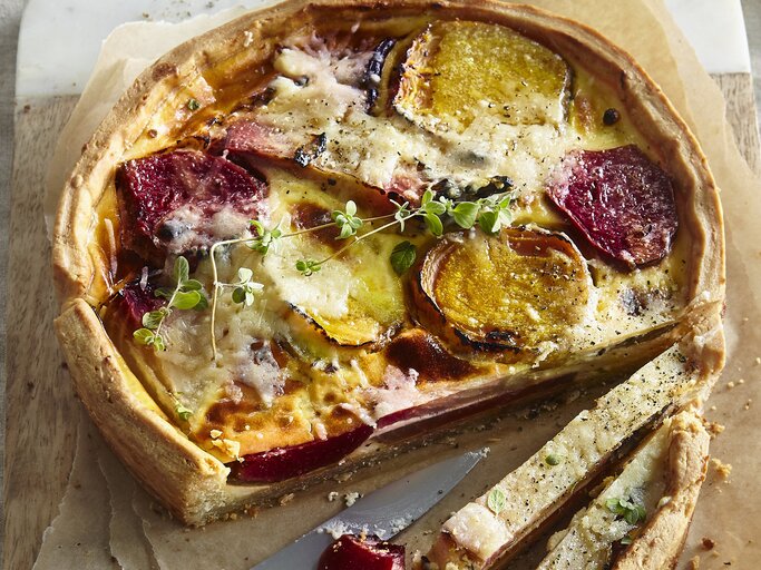 Angeschnittene Quiche mit Thymianzweig, geschmolzenem Käse auf Backpapier und geteiltem Holzbrett mit Steingutrand aus der Vogelperspektive. Rote Bete und Gelbe Bete in Scheiben sichtbar. Der Rand der Quiche leicht erhöht und gebräunt. Zwei Scheiben längs aufgeschnitten. Messer mit silberner Klinge und Holzgriff zwischen den Scheiben aufgelegt. | © StockFood / Profimedia
