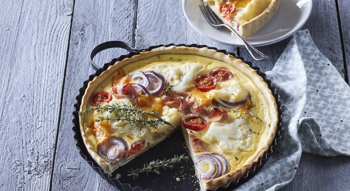 Eine Backform mit der frisch gebackenen Blumenkohl-Quiche mit Schinken und Räucherlachs neben einem Teller auf dem ein Stück der Quiche mit einer Gabel liegt, vor einem hellblauem Hintergrund.