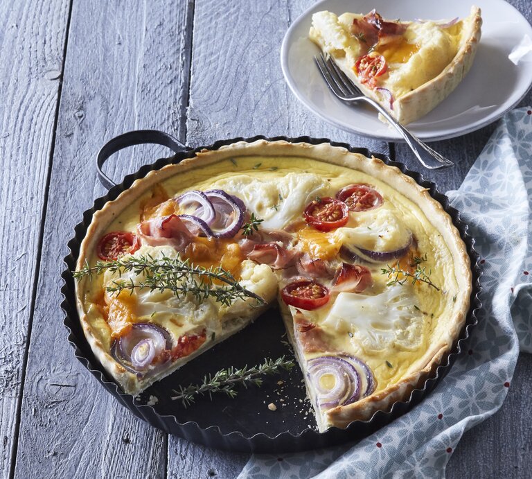 Eine Backform mit der frisch gebackenen Blumenkohl-Quiche mit Schinken und Räucherlachs neben einem Teller auf dem ein Stück der Quiche mit einer Gabel liegt, vor einem hellblauem Hintergrund. | © StockFood / Profimedia