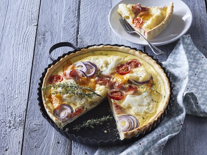 Eine Backform mit der frisch gebackenen Blumenkohl-Quiche mit Schinken und Räucherlachs neben einem Teller auf dem ein Stück der Quiche mit einer Gabel liegt, vor einem hellblauem Hintergrund. | © StockFood / Profimedia