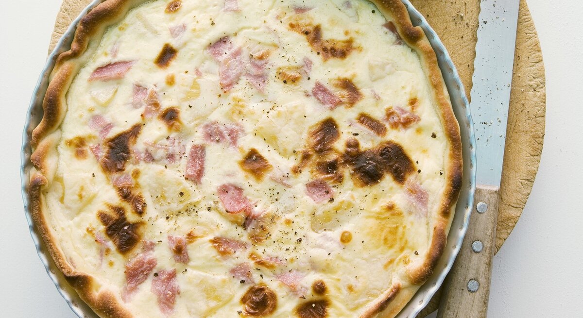 In einer runden Form ist eine Quiche Hawaii angerichtet. Sie ist mit Schinken und Ananas zubereitet.