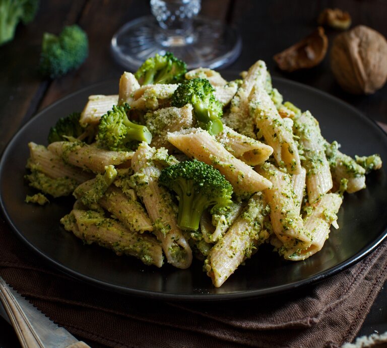 Ein Teller Brokkoli-Grünkohl-Pasta, daneben eine Gabel, im Hintergrund Walnüsse und Brokkoli. | © Adobe Stock/ katrinshine