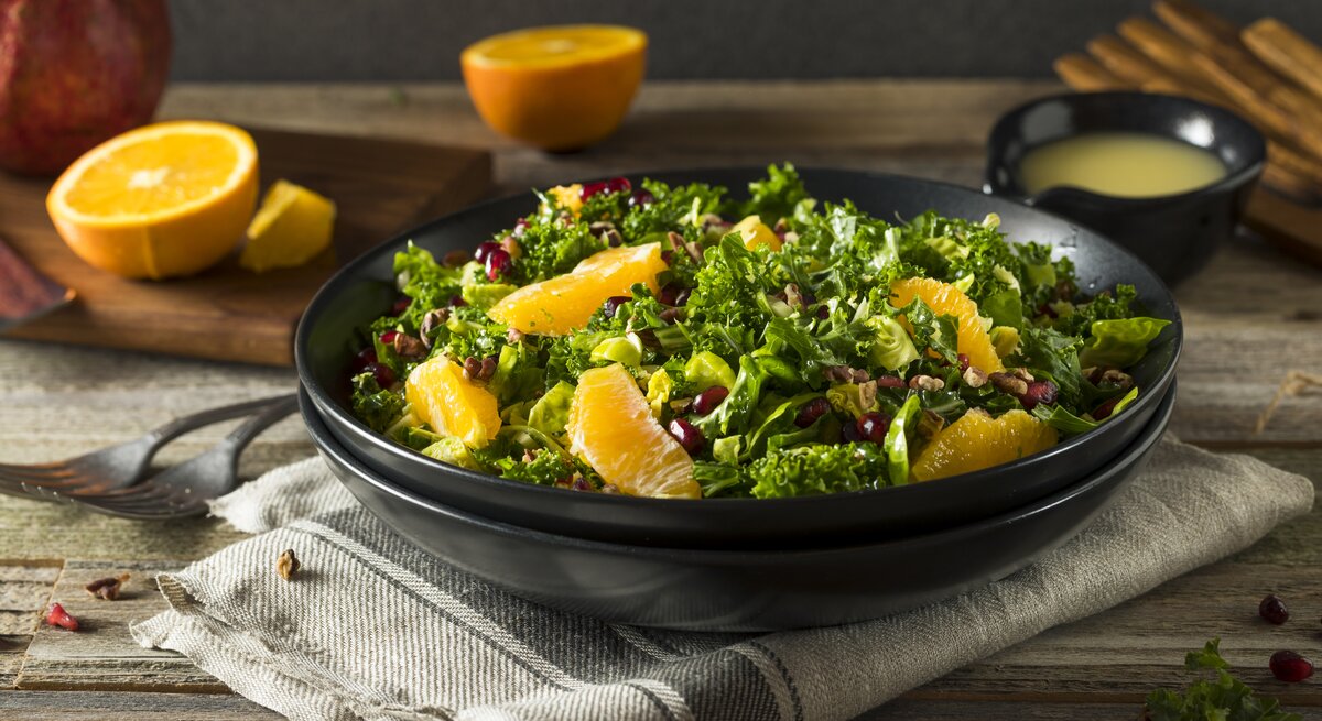 Auf einem Tisch steht ein schwarzer Teller. In dem ist ein Orangen-Grünkohl-Salat. Auf dem Tisch liegen Orangen-Hälften und ein Granatapfel.