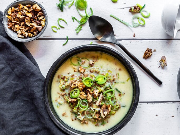 Eine schwarze Schüssel mit der Suppe auf einem weißen Tisch. Daneben ein Schälchen mit Walnüssen und geschnittenem Lauch. | © GettyImages/ Westend61