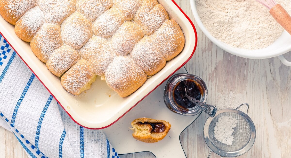 Draufsicht: Weißer Untergrung, auf einem Kuchenhandtuch eine Auflaufform mit selbst gemachten Hefe-Buchteln. Daneben eine Schüssel mit Mehl, ein geöffnetes Glas Fruchtaufstrich, ein Sieb mit Puderzucker und eine angebissene Buchtel.