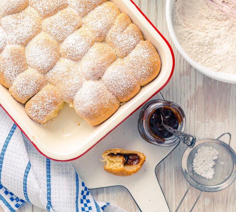 Draufsicht: Weißer Untergrung, auf einem Kuchenhandtuch eine Auflaufform mit selbst gemachten Hefe-Buchteln. Daneben eine Schüssel mit Mehl, ein geöffnetes Glas Fruchtaufstrich, ein Sieb mit Puderzucker und eine angebissene Buchtel. | © Shutterstock / Ingrid Balabanova