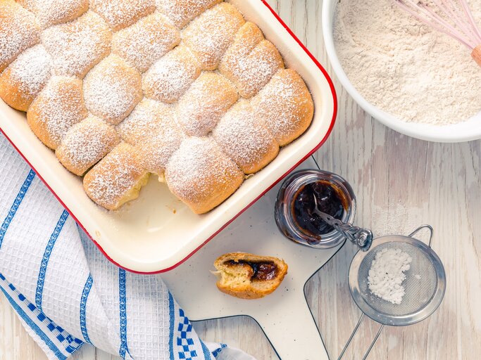 Draufsicht: Weißer Untergrung, auf einem Kuchenhandtuch eine Auflaufform mit selbst gemachten Hefe-Buchteln. Daneben eine Schüssel mit Mehl, ein geöffnetes Glas Fruchtaufstrich, ein Sieb mit Puderzucker und eine angebissene Buchtel. | © Shutterstock / Ingrid Balabanova