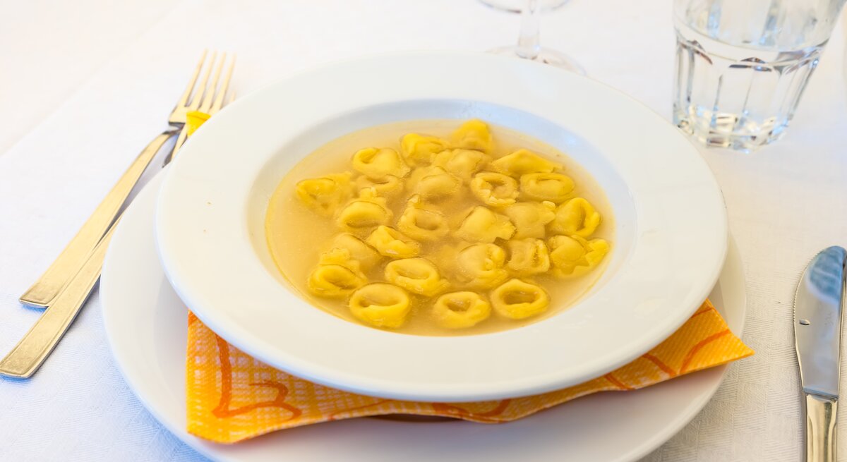 Teller mit klarer Brühe und Tortellini auf weißem Tischtuch. 
