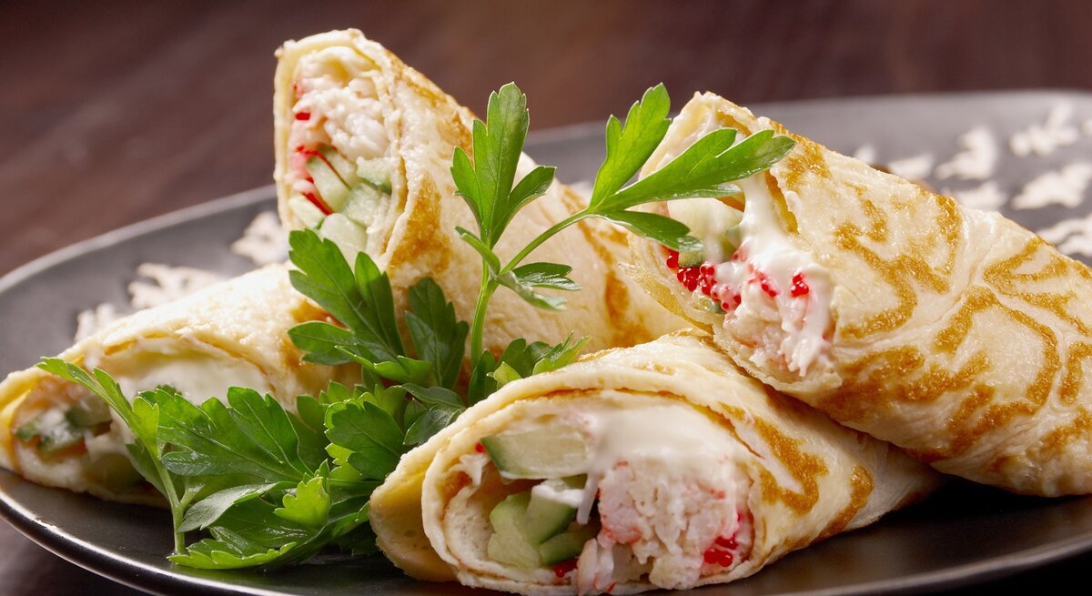 Vier angeschnittene Wraps mit Flusskrebs, Mayonnaise und Gurke auf einem grauen Teller und einem dunklen Holzuntergrund.