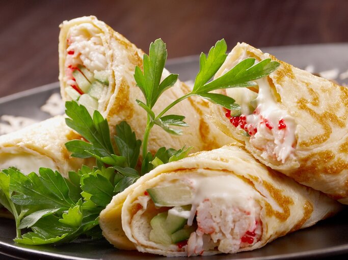 Vier angeschnittene Wraps mit Flusskrebs, Mayonnaise und Gurke auf einem grauen Teller und einem dunklen Holzuntergrund. | © Shutterstock/ Fanfo