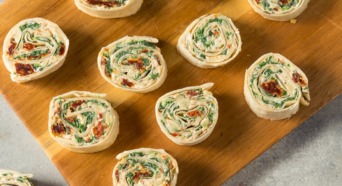 Wrap-Röllchen mit Feta, Spinat & getrockneten Tomaten auf Holzbrett. 