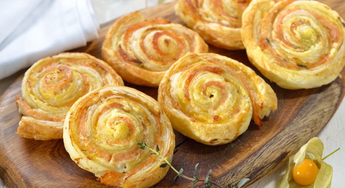 Sechs Blätterteigschnecken mit Lachs angerichtet auf einem Holzbrett. 