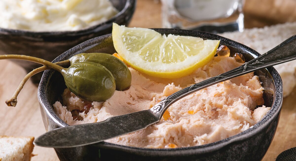 Frontalsicht: ein Räucherlachs-Dip mit Schmand in einemr schwarzen Schüssel. Darauf liegen Kapernäpfel und eine Zitronenscheibe. Im Hintergund stehen ein Schälchen Butter und ein Glas mit Räucherlachdip.
