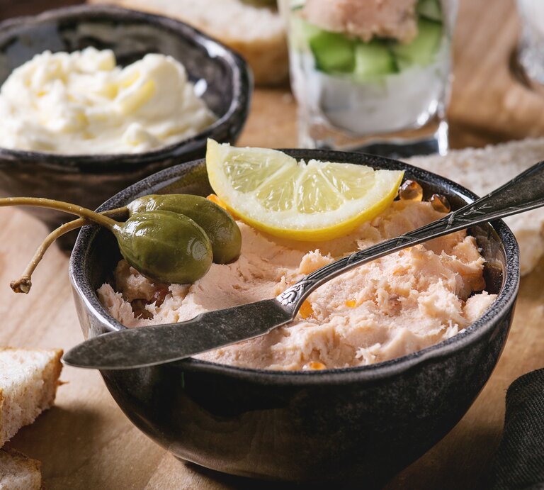 Frontalsicht: ein Räucherlachs-Dip mit Schmand in einemr schwarzen Schüssel. Darauf liegen Kapernäpfel und eine Zitronenscheibe. Im Hintergund stehen ein Schälchen Butter und ein Glas mit Räucherlachdip. | © Shutterstock / Natasha Breen
