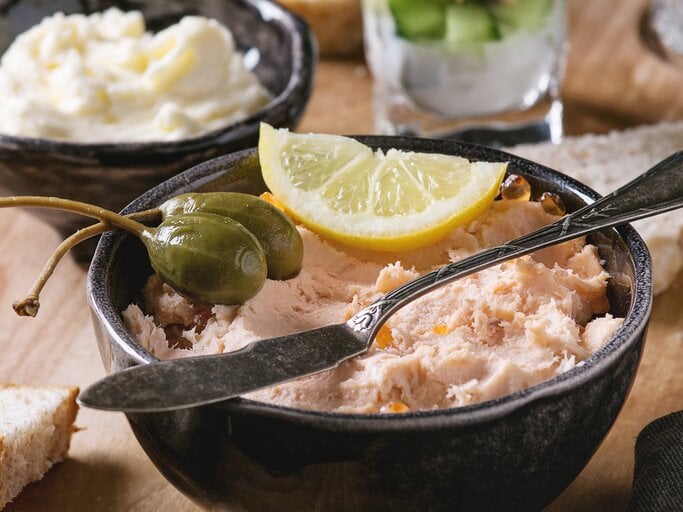 Frontalsicht: ein Räucherlachs-Dip mit Schmand in einemr schwarzen Schüssel. Darauf liegen Kapernäpfel und eine Zitronenscheibe. Im Hintergund stehen ein Schälchen Butter und ein Glas mit Räucherlachdip. | © Shutterstock / Natasha Breen