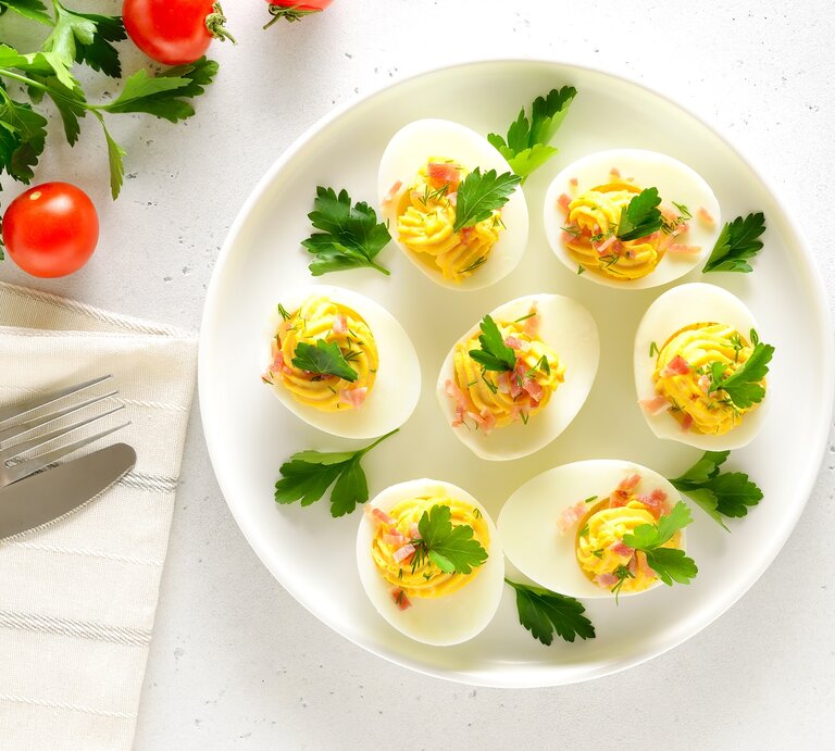 Draufsicht: Ein weißer Untergrund. Gefüllte Eier mit Speck auf einem weißen Teller. Daneben sind Tomaten, frische Petersilie und ein Besteck. | © © Shutterstock / Nadezhda Nesterova