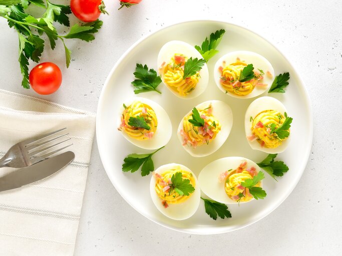 Draufsicht: Ein weißer Untergrund. Gefüllte Eier mit Speck auf einem weißen Teller. Daneben sind Tomaten, frische Petersilie und ein Besteck. | © © Shutterstock / Nadezhda Nesterova