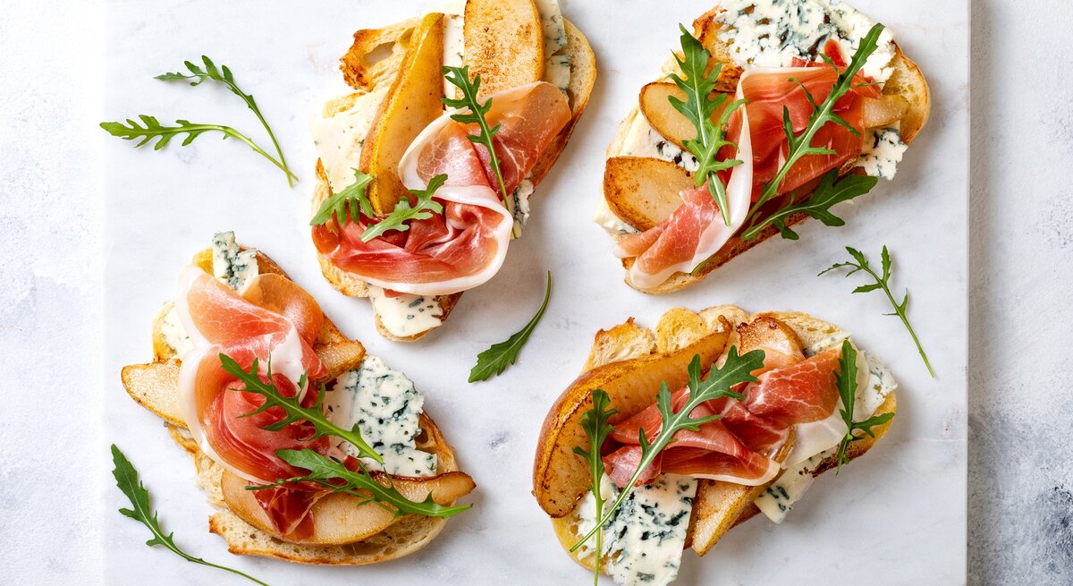 Crostini mit Birne, Schinken und Gorgonzola auf einer grauen Keramik-Platte. 