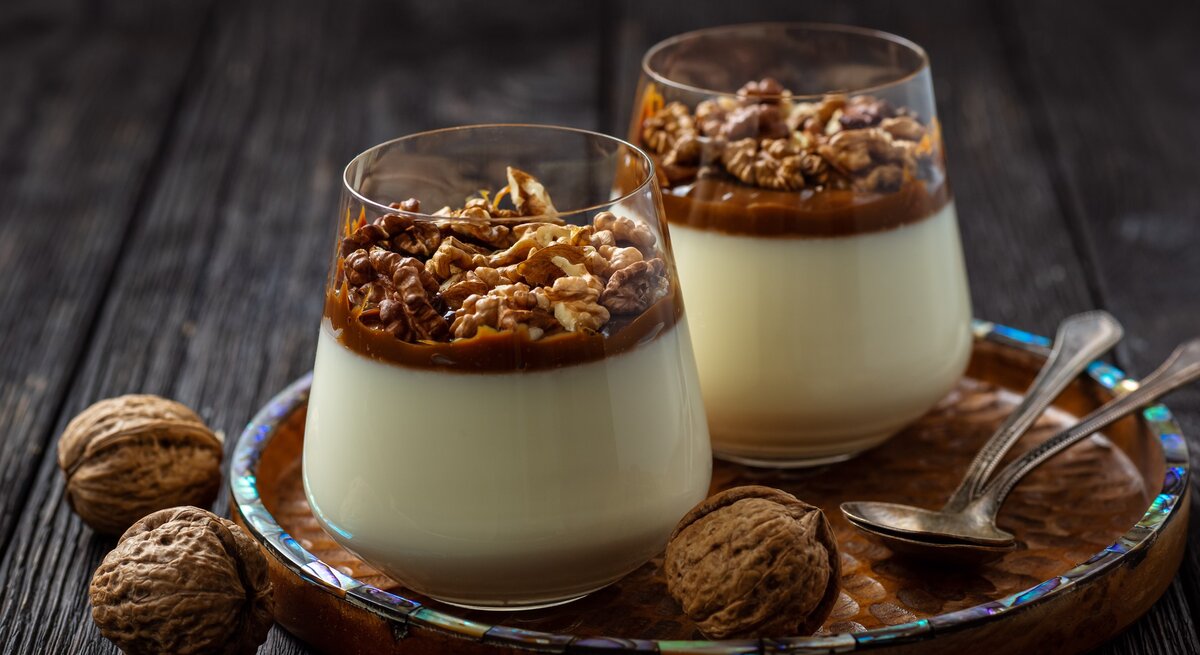 Weihnachtliches Panna Cotta mit Gewürzkaramell und Walnüssen im konischen Glas auf Holzteller mit Perlmuttrand auf dunklem Holztisch in der Frontalansicht. Ganze Walnüsse und zweit Silberlöffel als Dekoration.