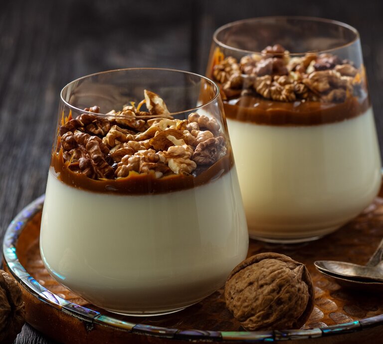Weihnachtliches Panna Cotta mit Gewürzkaramell und Walnüssen im konischen Glas auf Holzteller mit Perlmuttrand auf dunklem Holztisch in der Frontalansicht. Ganze Walnüsse und zweit Silberlöffel als Dekoration. | © Oleg Bannikov