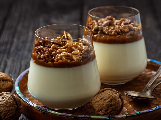 Weihnachtliches Panna Cotta mit Gewürzkaramell und Walnüssen im konischen Glas auf Holzteller mit Perlmuttrand auf dunklem Holztisch in der Frontalansicht. Ganze Walnüsse und zweit Silberlöffel als Dekoration. | © Oleg Bannikov