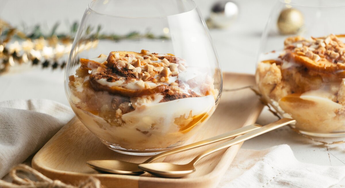 Bratapfel-Dessert im Glas mit Vanillesoße, Gewürzkaramell, Keksen und Mandelsplittern auf Holzteller, beiger Stoffserviette und Tisch mit weißem Tischtuch. Ein zweites Glas im Hintergrund angedeutet. Weihnachtdekoration in grün, gold, silber und Schwarz im Hintergrund unscharf. Aufnahme aus der Frontale.