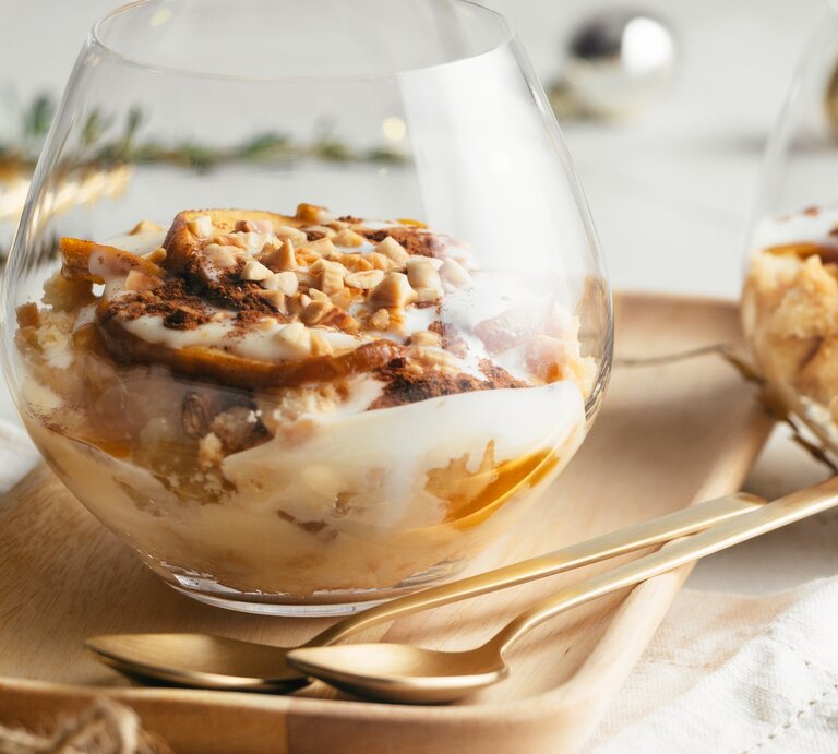 Bratapfel-Dessert im Glas mit Vanillesoße, Gewürzkaramell, Keksen und Mandelsplittern auf Holzteller, beiger Stoffserviette und Tisch mit weißem Tischtuch. Ein zweites Glas im Hintergrund angedeutet. Weihnachtdekoration in grün, gold, silber und Schwarz im Hintergrund unscharf. Aufnahme aus der Frontale. | © ASIFE