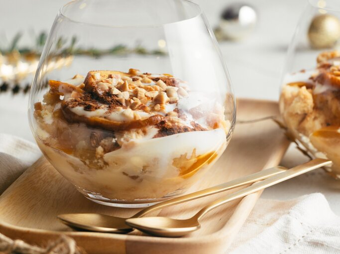 Bratapfel-Dessert im Glas mit Vanillesoße, Gewürzkaramell, Keksen und Mandelsplittern auf Holzteller, beiger Stoffserviette und Tisch mit weißem Tischtuch. Ein zweites Glas im Hintergrund angedeutet. Weihnachtdekoration in grün, gold, silber und Schwarz im Hintergrund unscharf. Aufnahme aus der Frontale. | © ASIFE
