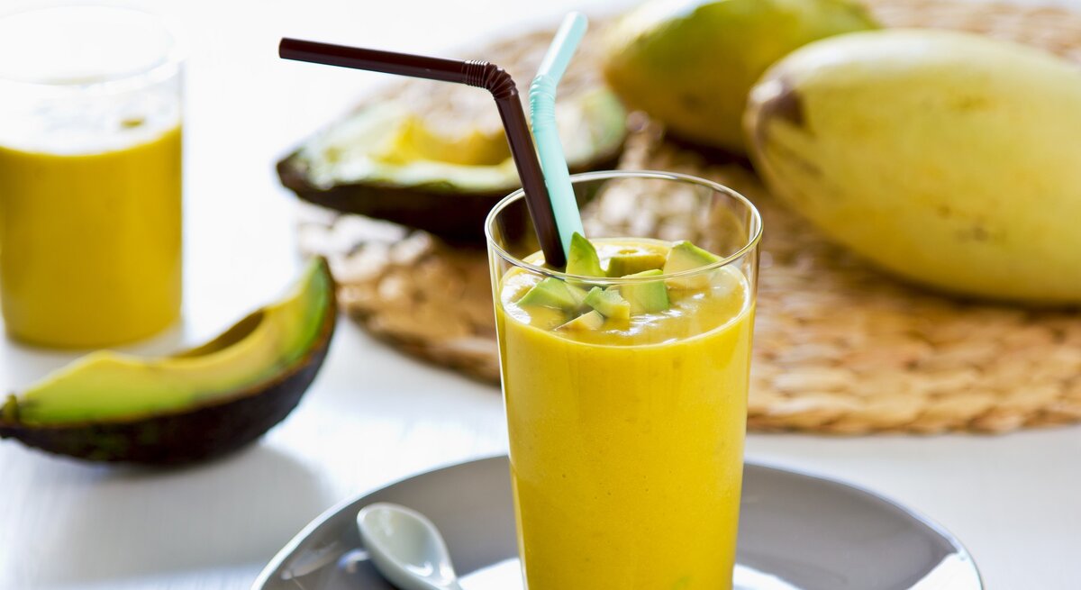 Ein Glas mit dem gelben Smoothie auf einem hellen Tisch mit Avocado und Mango im Hintergrund.