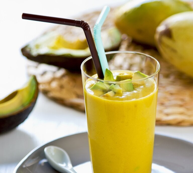 Ein Glas mit dem gelben Smoothie auf einem hellen Tisch mit Avocado und Mango im Hintergrund. | © Shutterstock/ vanillaechoes 