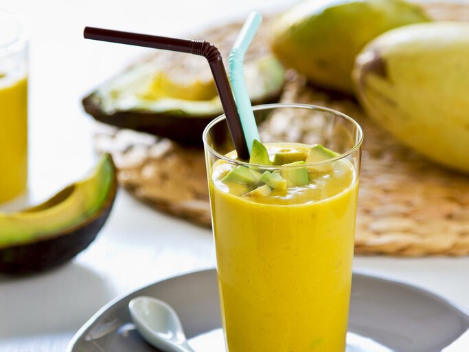 Ein Glas mit dem gelben Smoothie auf einem hellen Tisch mit Avocado und Mango im Hintergrund. | © Shutterstock/ vanillaechoes 