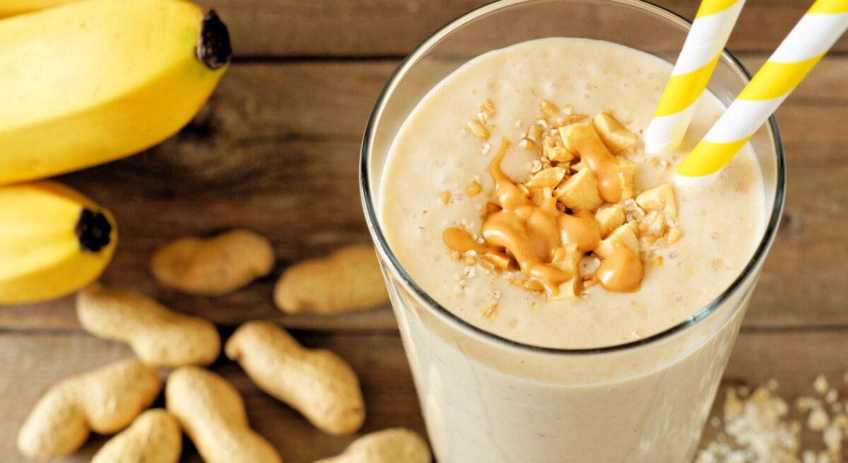 Erdnuss-Bananen-Shake mit Haferflocken im Glas mit Strohhalm und Toppings auf Holztisch. Daneben Erdnüsse und Bananen. Draufsicht.