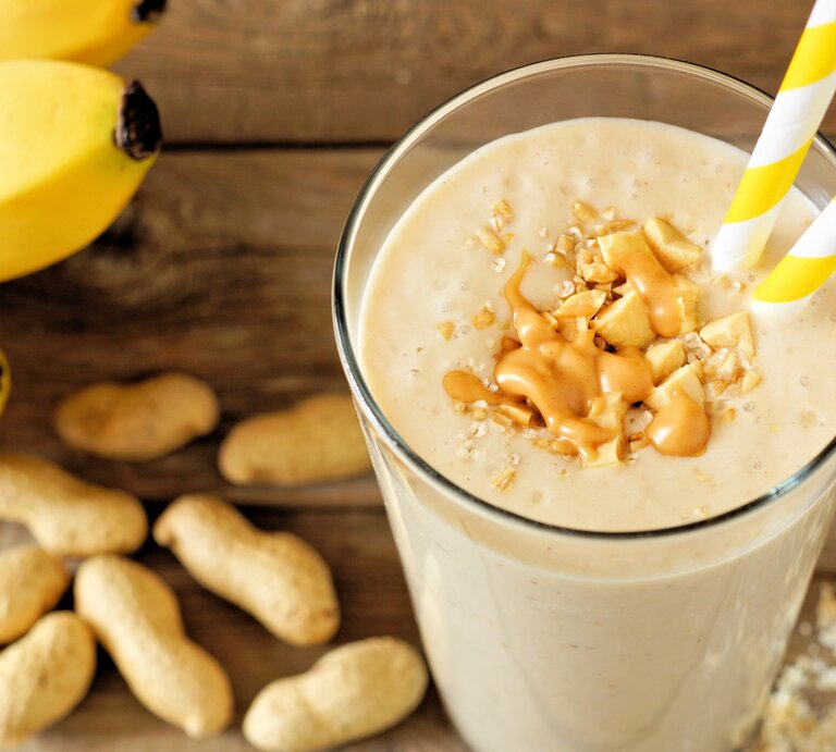 Erdnuss-Bananen-Shake mit Haferflocken im Glas mit Strohhalm und Toppings auf Holztisch. Daneben Erdnüsse und Bananen. Draufsicht. | © Getty Images / jenifoto