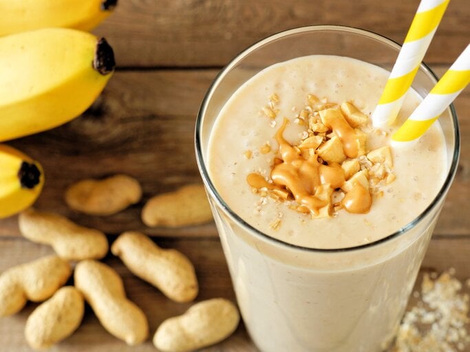 Erdnuss-Bananen-Shake mit Haferflocken im Glas mit Strohhalm und Toppings auf Holztisch. Daneben Erdnüsse und Bananen. Draufsicht. | © Getty Images / jenifoto