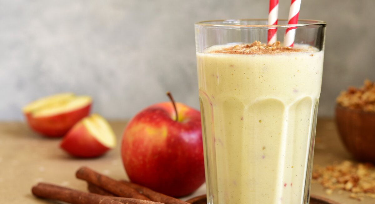 Ein Glas mit dem Smoothie auf einem hellbraunem Untergrund. Daneben ein paar Zimtstangen und ein paar Apfelspalten.
