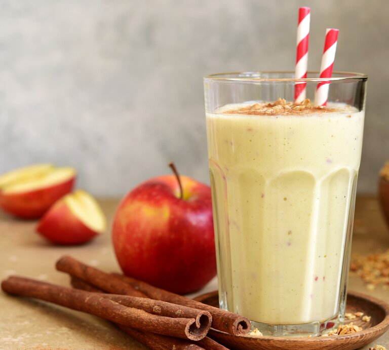 Ein Glas mit dem Smoothie auf einem hellbraunem Untergrund. Daneben ein paar Zimtstangen und ein paar Apfelspalten. | © Shutterstock/ Liliya Kandrashevich