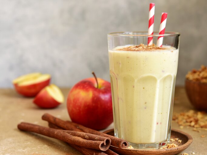 Ein Glas mit dem Smoothie auf einem hellbraunem Untergrund. Daneben ein paar Zimtstangen und ein paar Apfelspalten. | © Shutterstock/ Liliya Kandrashevich