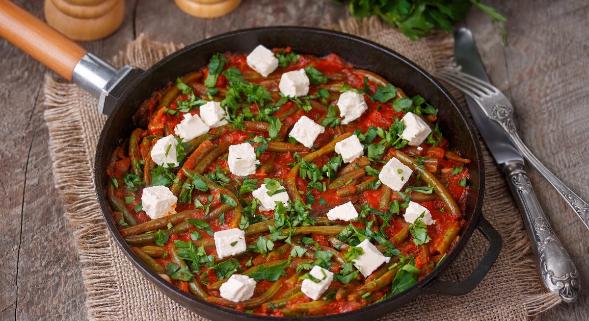 Draufsicht, schräg: eine Grüne Bohne-Pfanne mit Feta auf einem Holztisch und einem Tuch aus Leinen. Daneben liegen ein Besteck und frische Petersilie.