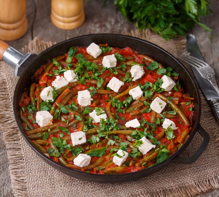Draufsicht, schräg: eine Grüne Bohne-Pfanne mit Feta auf einem Holztisch und einem Tuch aus Leinen. Daneben liegen ein Besteck und frische Petersilie. | © Shutterstock / Dementieva Iryna