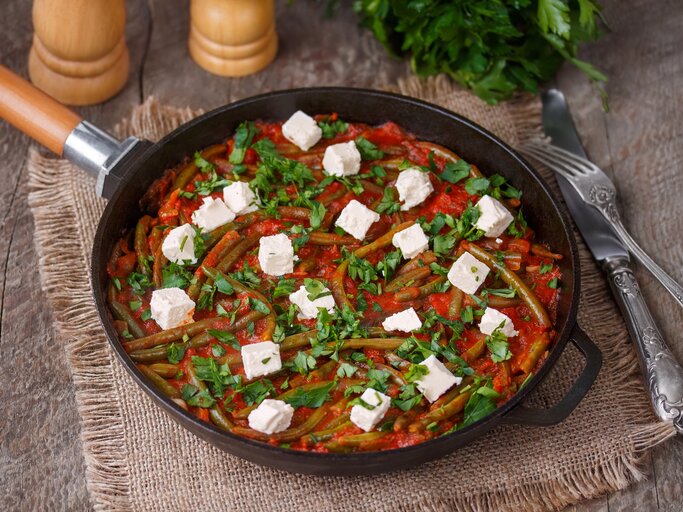 Draufsicht, schräg: eine Grüne Bohne-Pfanne mit Feta auf einem Holztisch und einem Tuch aus Leinen. Daneben liegen ein Besteck und frische Petersilie. | © Shutterstock / Dementieva Iryna