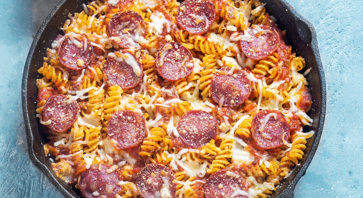 Spirelli-Salami-Pfanne mit Tomaten in Gusspfanne auf blauem Untergrund. Oben im Bild rot-weiß-gestreiftes Tuch. Aufnahme aus der Vogelperspektive.