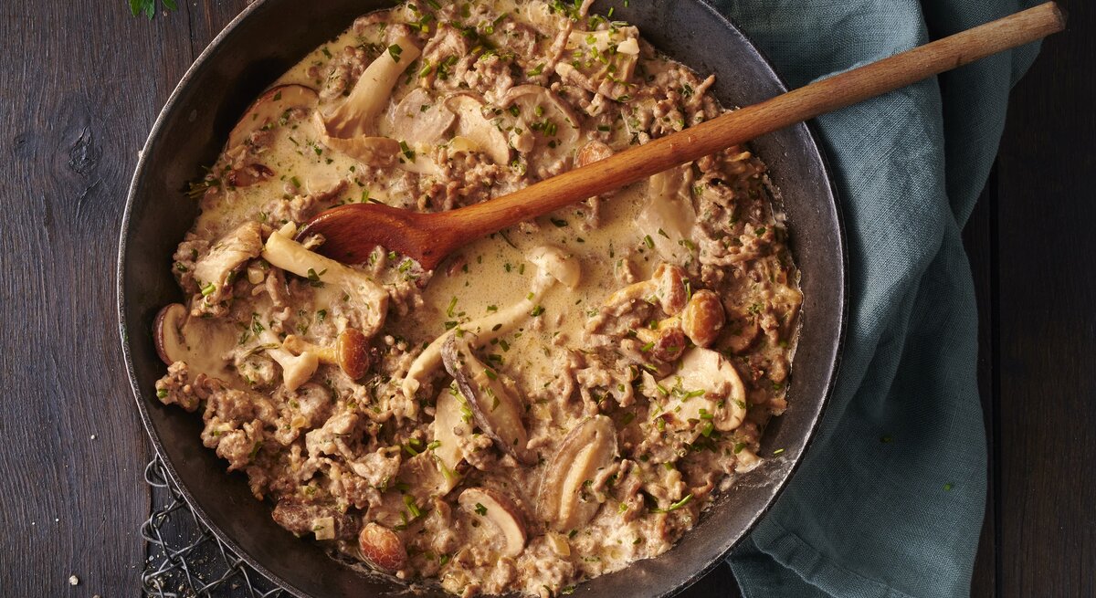Hackfleisch-Jägerpfanne mit  gemischten Pilzen und frischen Kräutern. Das Gericht befindet sich in einer rustikalen Pfanne und besteht aus gebräuntem Hackfleisch gemischt mit einer Vielzahl von Pilzen, die in verschiedenen Größen und Formen zu sehen sind. Die Pilze sind goldbraun geröstet.