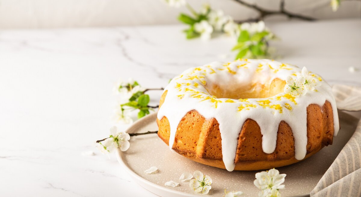 Ein Aperol-Gugelhupf auf einem runden Teller, der Kuchen mit Zuckerguss und Zitronenabrieb dekoriert, im Hintergrund Zweige und Blüten.