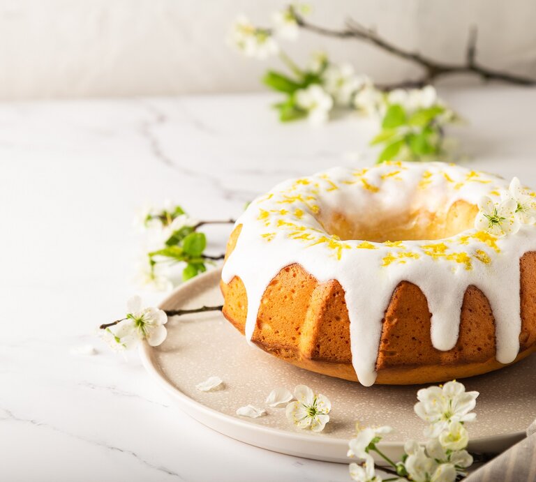 Ein Aperol-Gugelhupf auf einem runden Teller, der Kuchen mit Zuckerguss und Zitronenabrieb dekoriert, im Hintergrund Zweige und Blüten. | © Adobe Stock/ Anastasia Izofatova