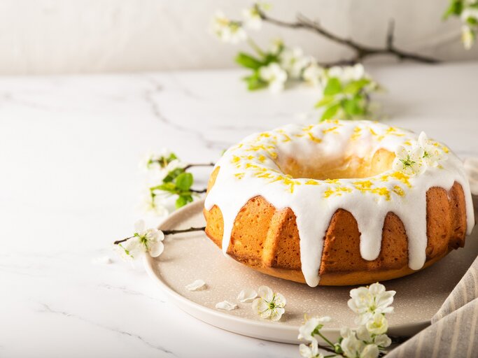 Ein Aperol-Gugelhupf auf einem runden Teller, der Kuchen mit Zuckerguss und Zitronenabrieb dekoriert, im Hintergrund Zweige und Blüten. | © Adobe Stock/ Anastasia Izofatova