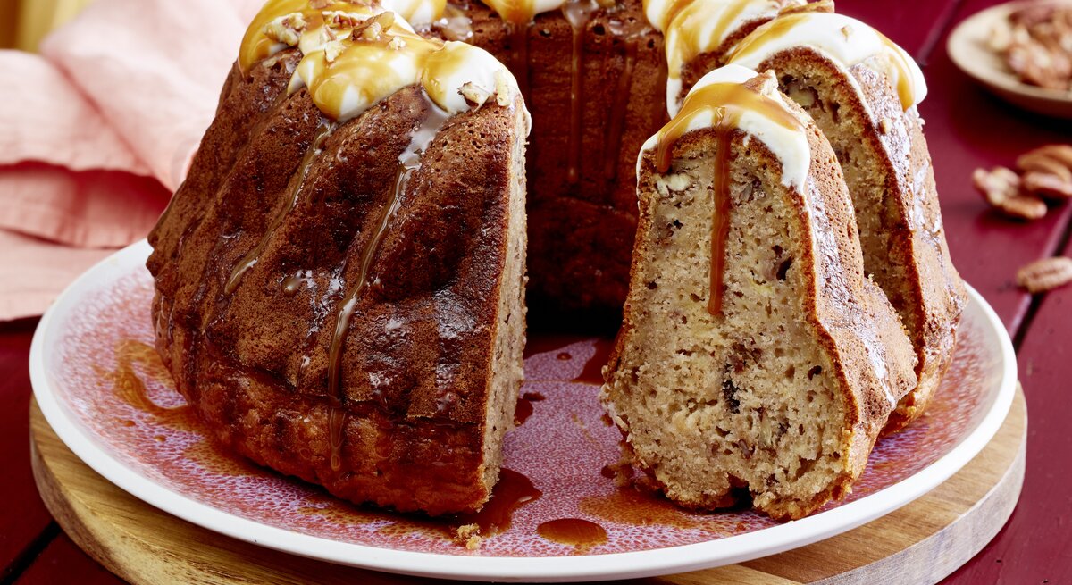 Eine Kuchenplatte mit dem angeschnittenen Apfel-Bananen-Gugelhupf mit Karamell-Soße übergossen und Pekan-Nüssen überstreut vor einem dunkel-roten Hintergrund.