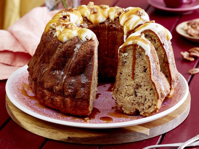 Eine Kuchenplatte mit dem angeschnittenen Apfel-Bananen-Gugelhupf mit Karamell-Soße übergossen und Pekan-Nüssen überstreut vor einem dunkel-roten Hintergrund. | © EAT CLUB/Isabell Triemer 