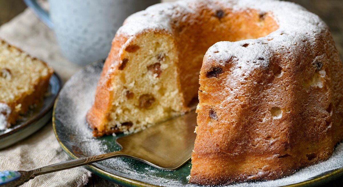 Frontalansicht: Ein angeschnittener Hefe-Gugelhupf steht auf einem Teller. Dahinter stehe eine Tasse und ein Stückchen Kuchen.