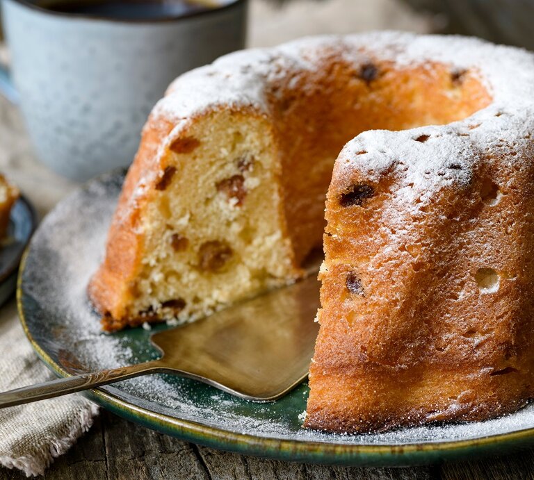 Frontalansicht: Ein angeschnittener Hefe-Gugelhupf steht auf einem Teller. Dahinter stehe eine Tasse und ein Stückchen Kuchen. | © Shutterstock / Stramp